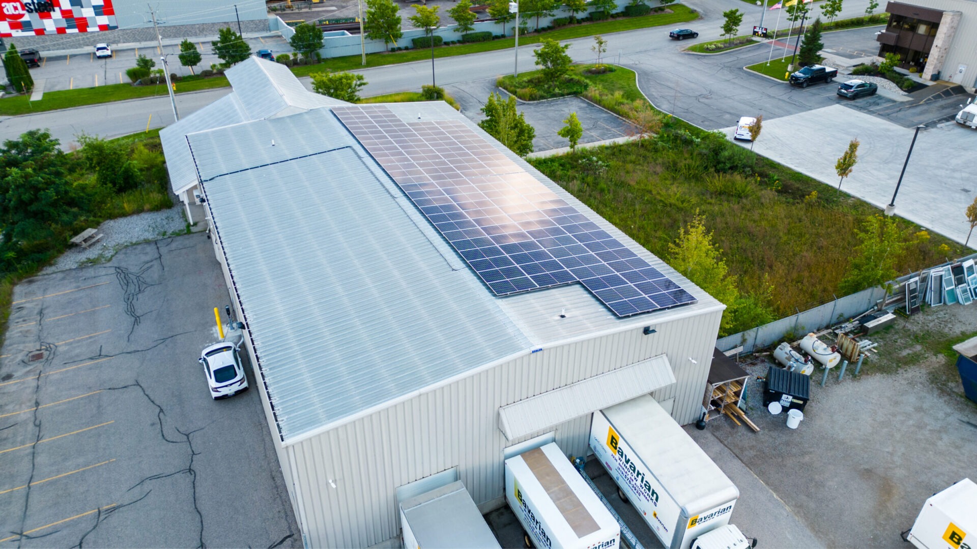 An industrial building with solar panels on its roof, surrounded by parking areas and greenery. Several trucks are parked beside the building.