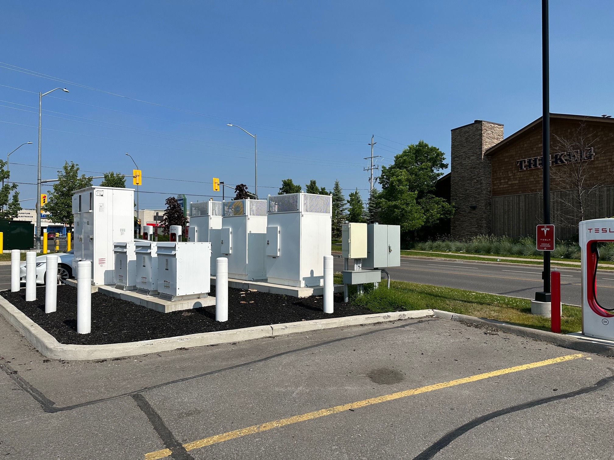 A Tesla charging station is near a road with traffic lights, adjacent to a building with "The Keg" signage, surrounded by greenery.