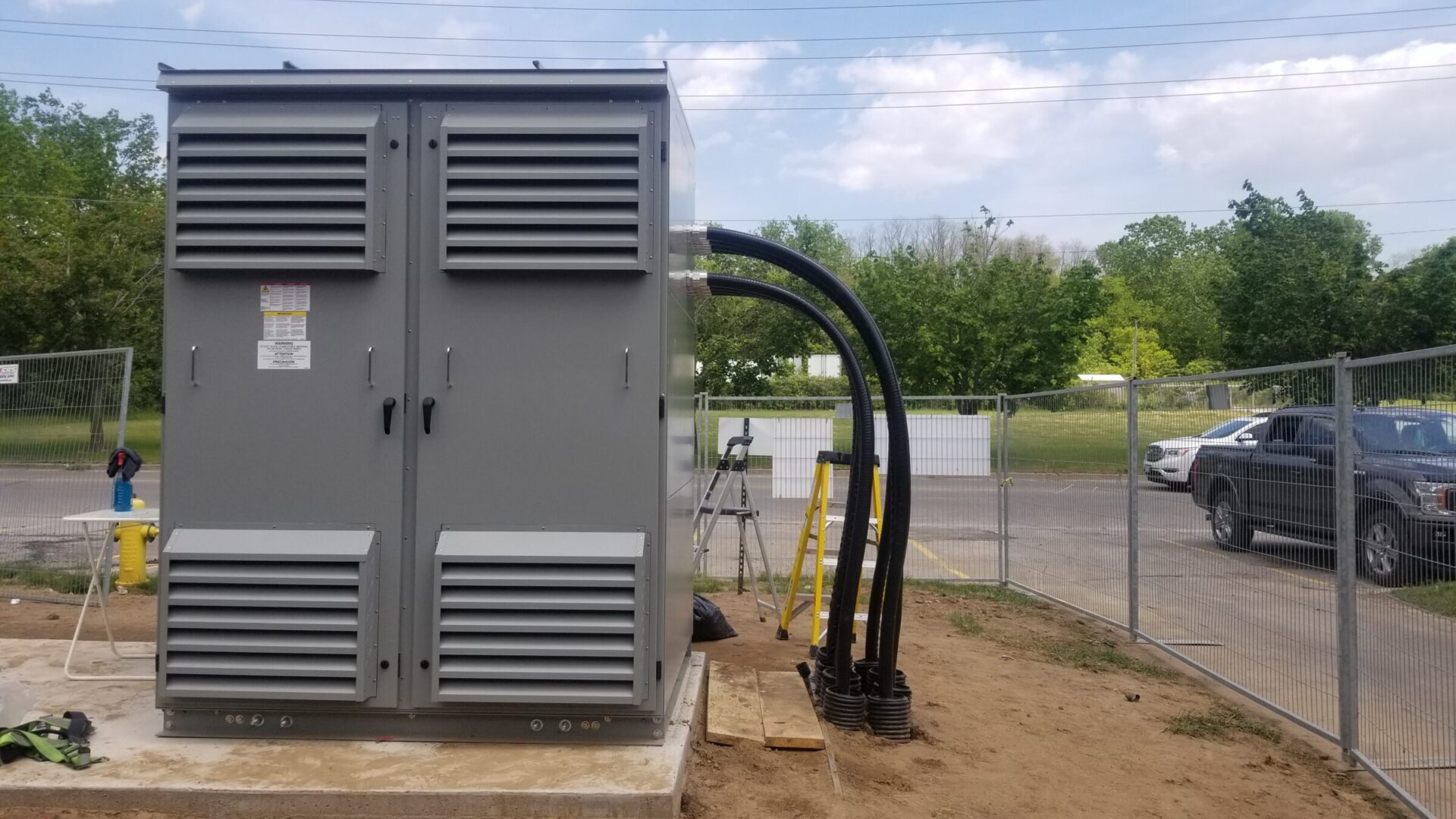 A large gray electrical transformer sits inside a fenced area. Thick black cables extend from it. Trees and a parked car are visible.