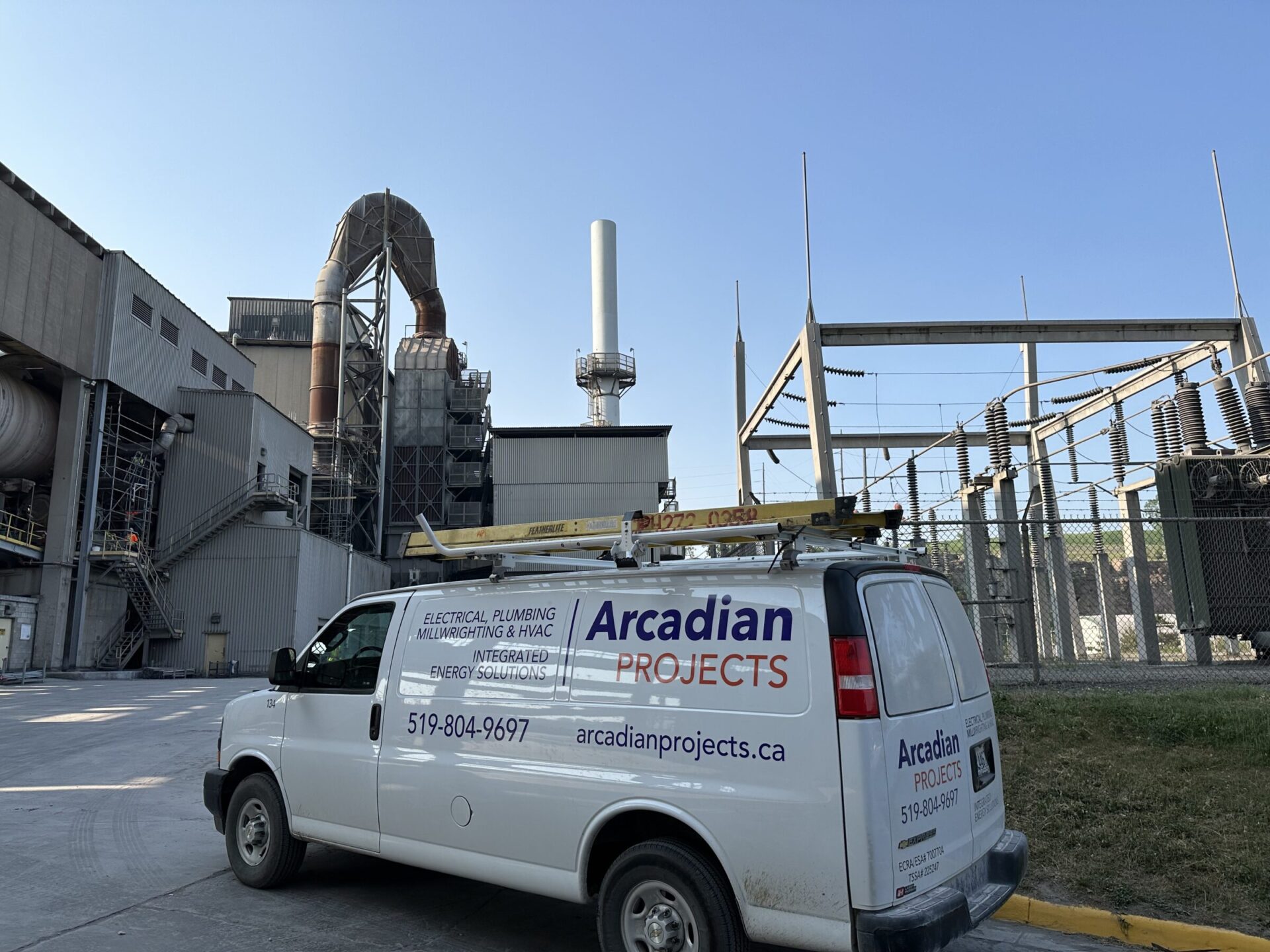 A white service van labeled "Arcadian Projects" is parked near an industrial facility with large machinery and electrical infrastructure. Clear blue sky.