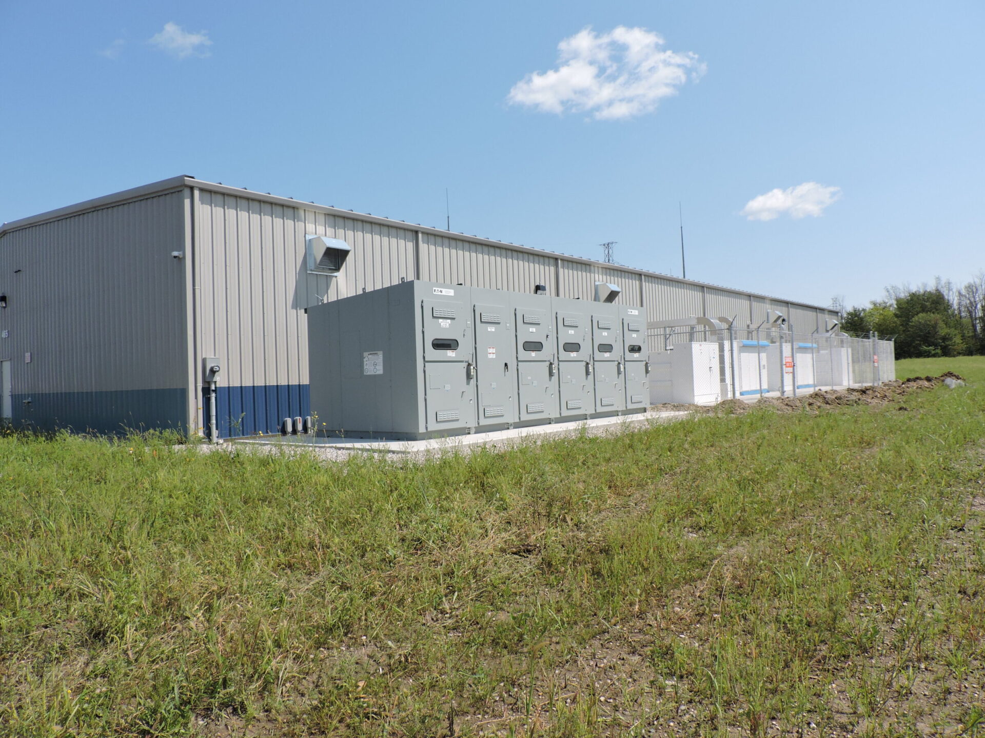 A large industrial building with electrical equipment in front, surrounded by grassy area under a clear blue sky with scattered clouds.