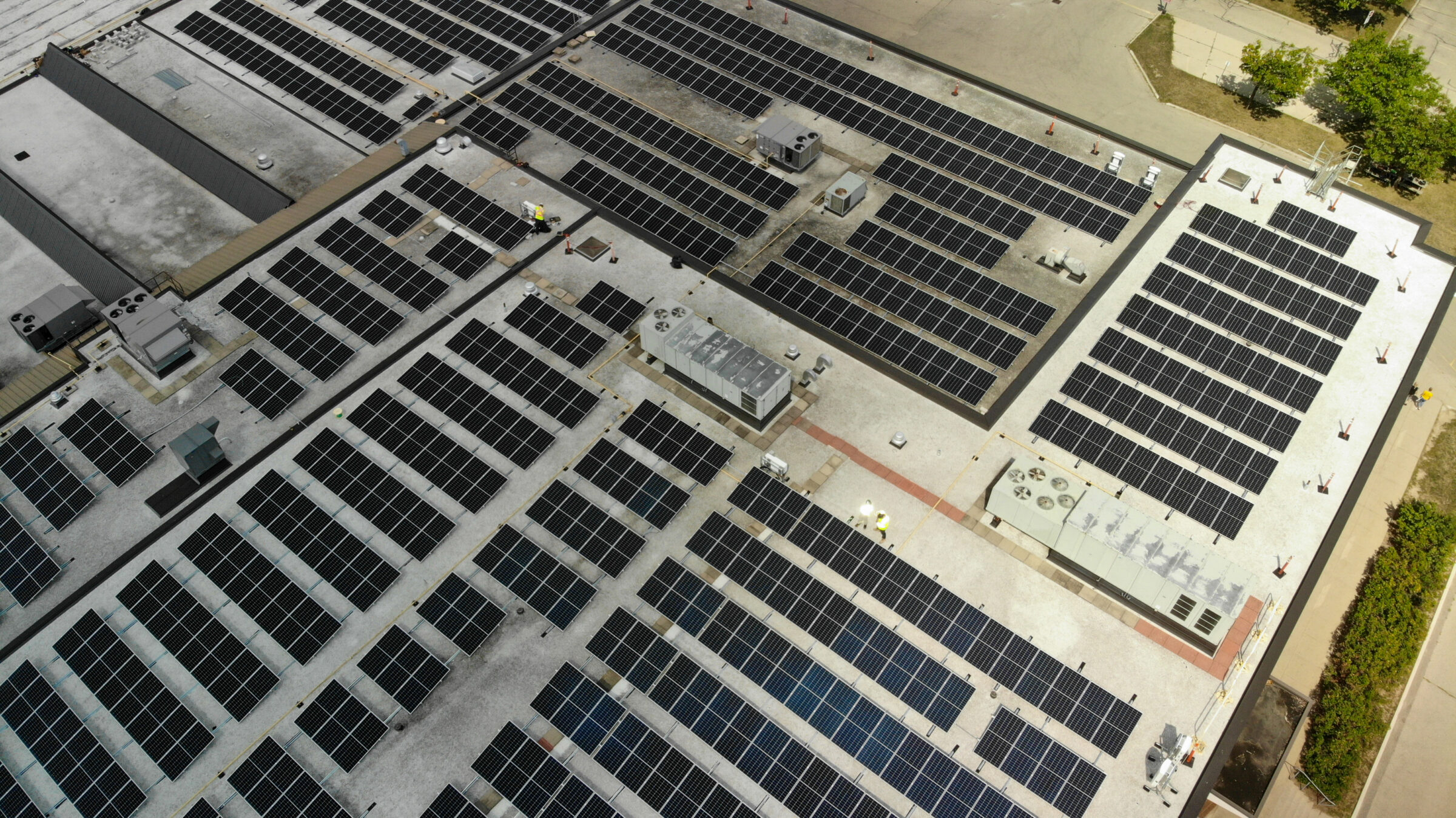 Aerial view of a rooftop covered with solar panels. Several people are visible, inspecting the installation. Nearby, trees and pathways are present.