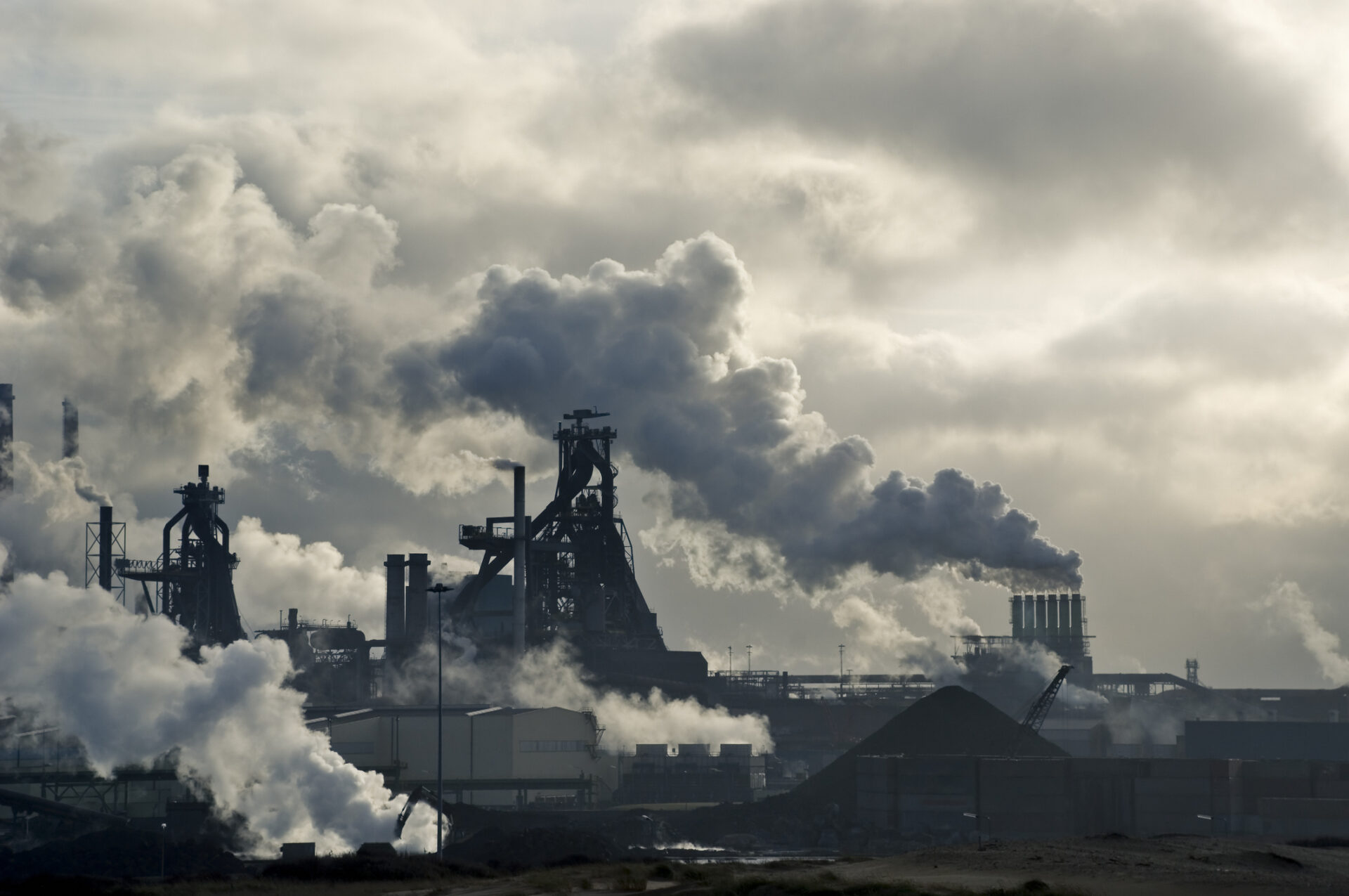 Industrial landscape with large smokestacks emitting clouds of smoke, contrasting against a cloudy sky. No recognizable landmarks or historical buildings are visible.