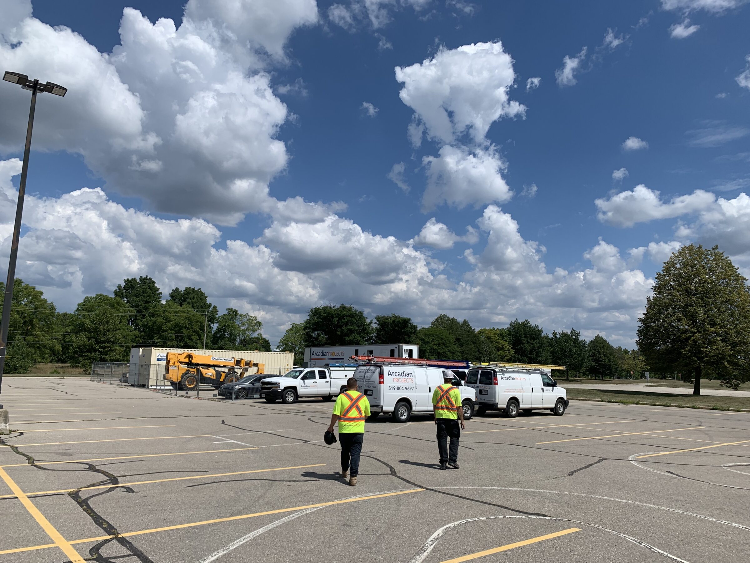 Two people wearing safety vests walk in a parking lot with parked trucks and construction equipment, surrounded by trees under a partly cloudy sky.