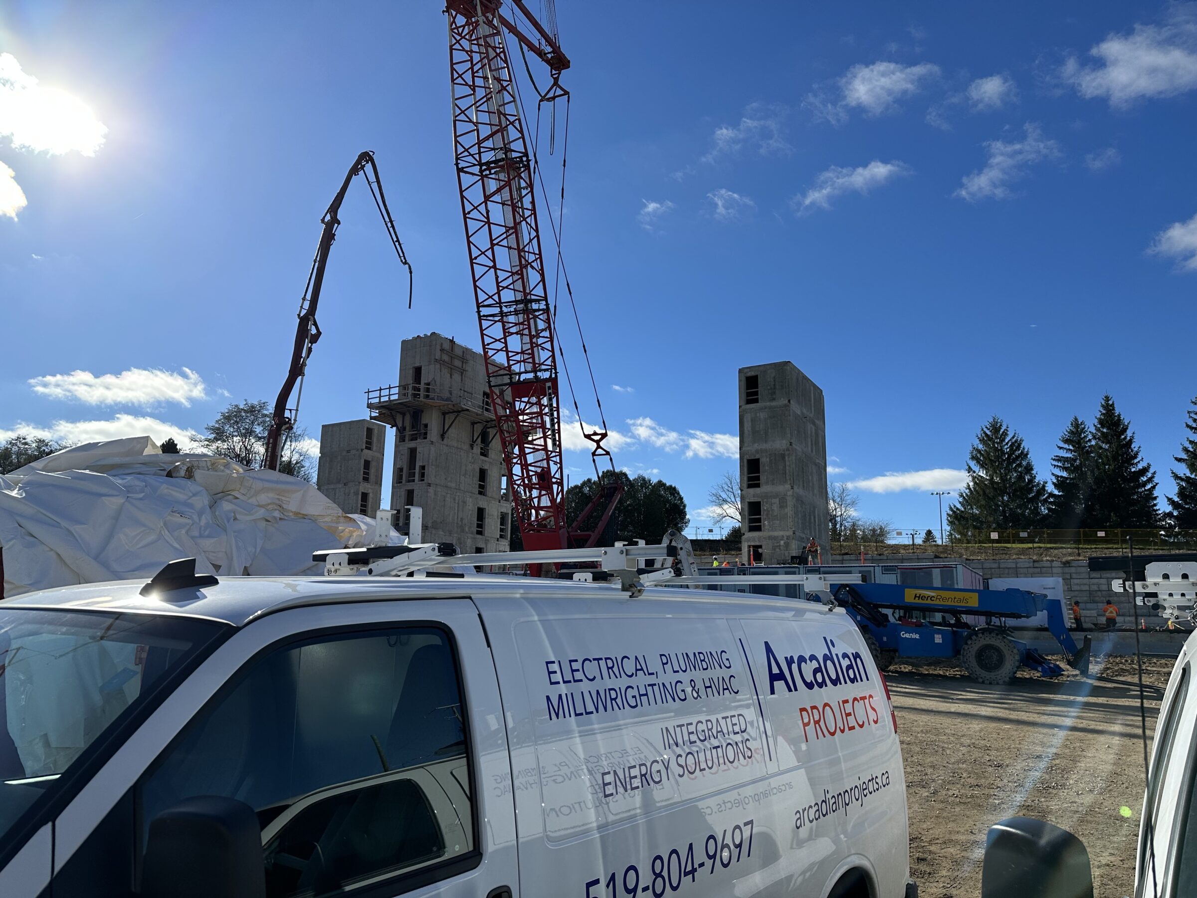 Construction site with cranes and unfinished buildings under a clear sky, featuring a van labeled "Arcadian Projects" in the foreground.