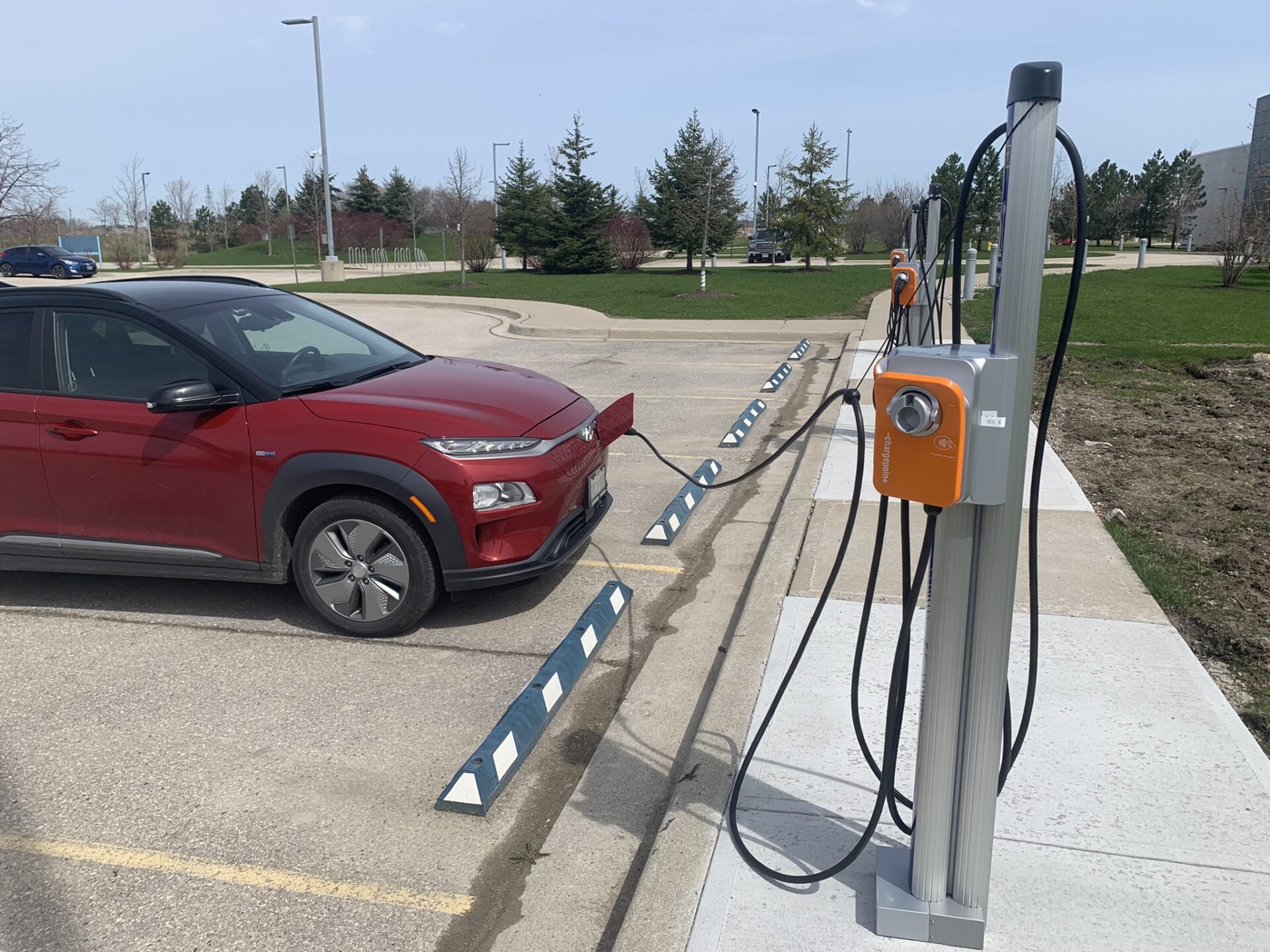 A red electric vehicle is parked beside charging stations in a parking lot with greenery and cloudy skies in the background.