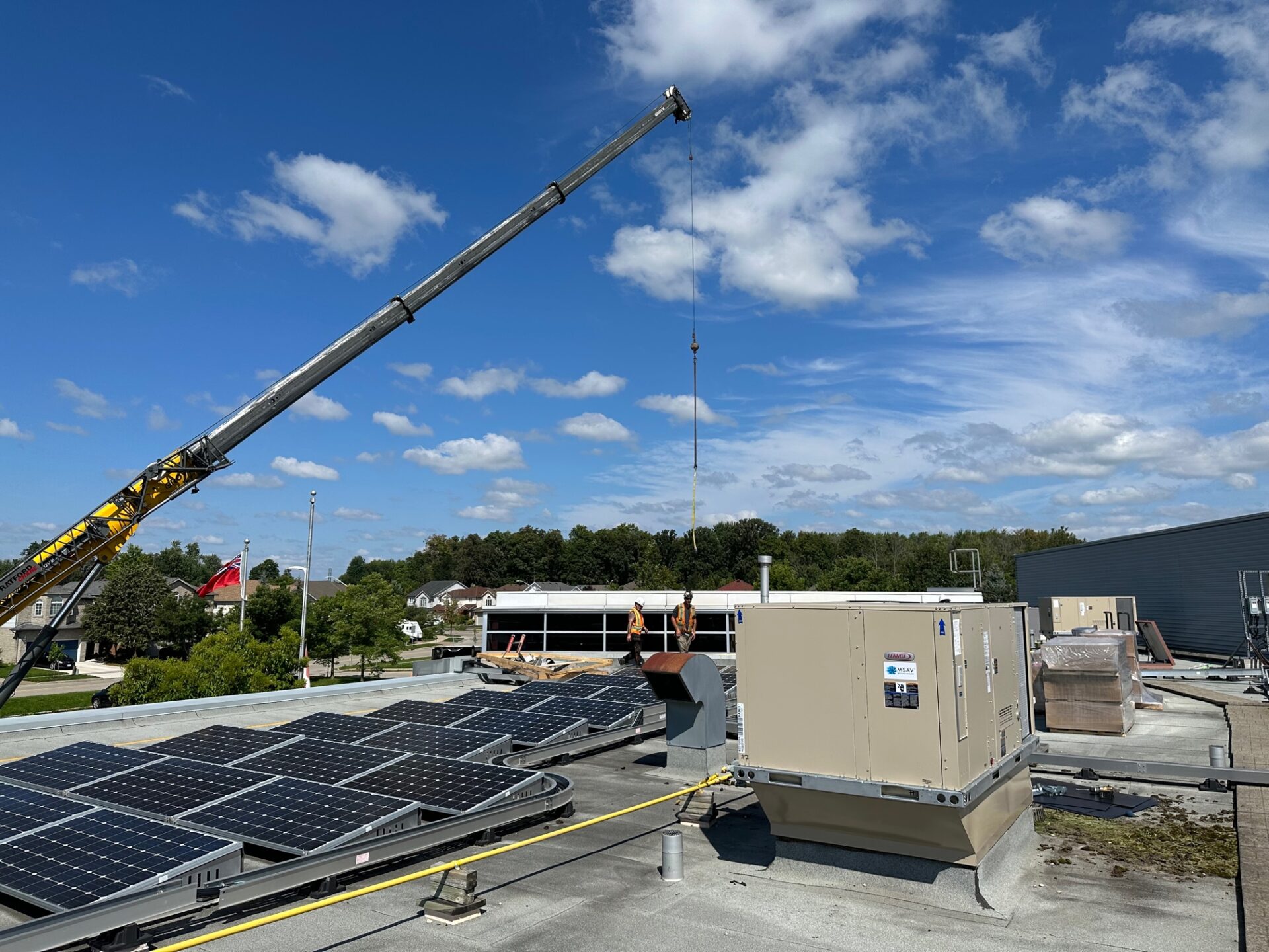 A rooftop with solar panels, a person operating a crane, and trees in the background under a partly cloudy sky.