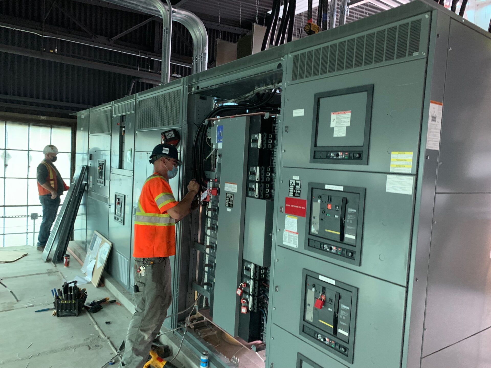 Two people in safety attire work on large electrical panels in an industrial setting. Various tools are visible on the floor.