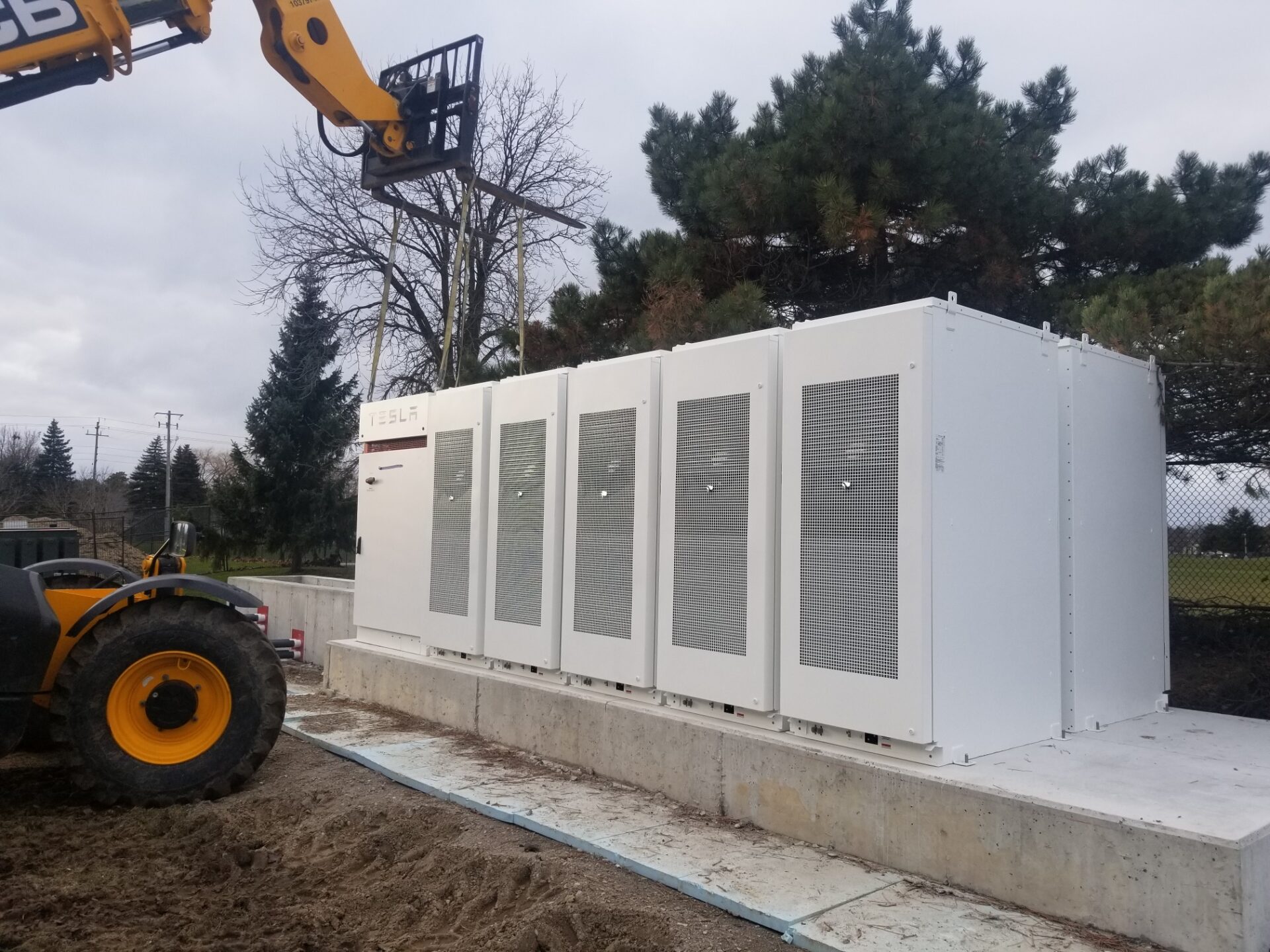 The image shows a Tesla battery storage unit installed outdoors, with a machine nearby. Trees and fencing are in the background under a cloudy sky.