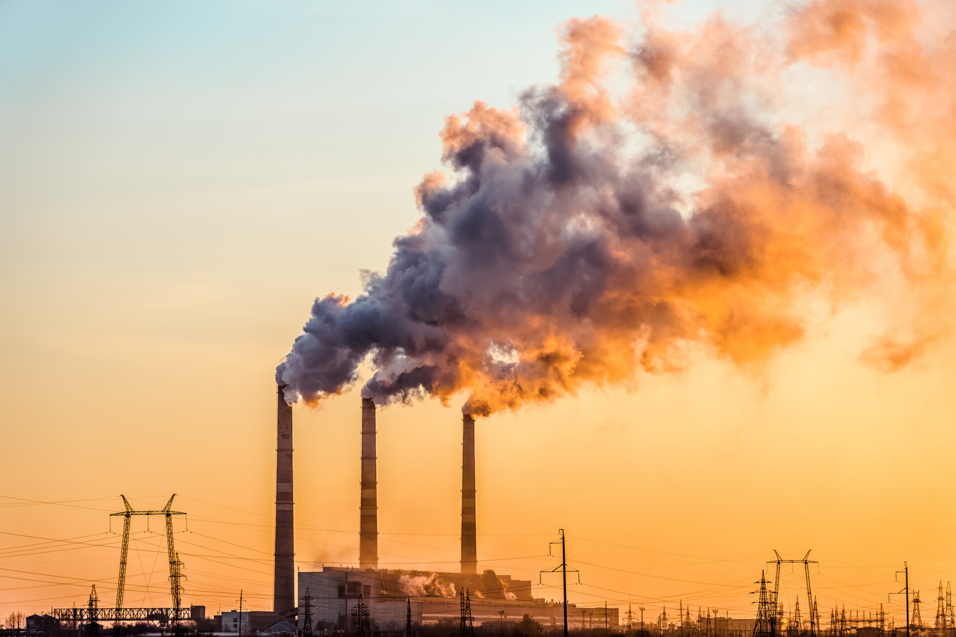 Three smokestacks release emissions against a vivid, colorful sunset sky. Power lines crisscross the industrial landscape, highlighting environmental contrasts and infrastructure.