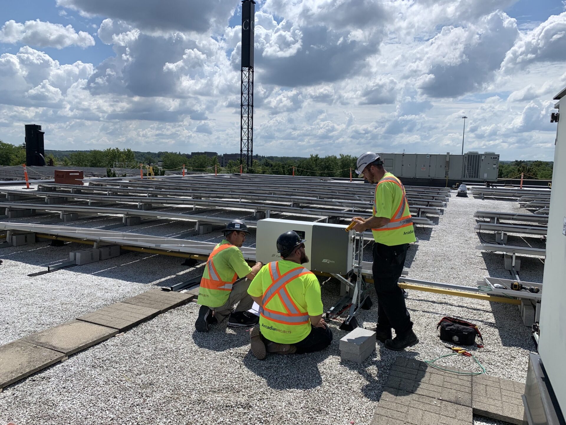 Three people in safety vests work on rooftop solar panels under a partly cloudy sky. Nearby equipment and trees in the background.