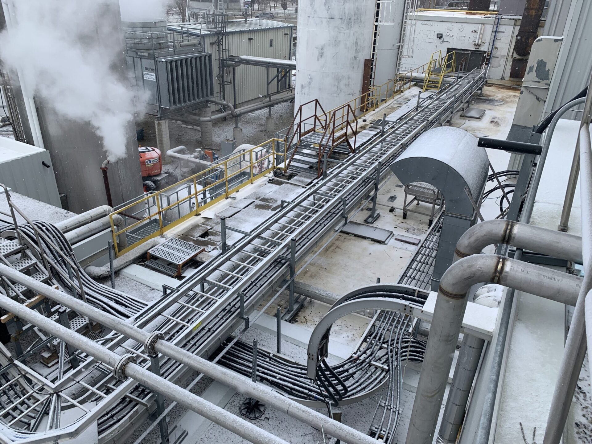 Industrial plant with extensive piping and metal walkways, emitting steam. Snow covers parts of the structure, creating a stark, mechanical atmosphere. No people visible.