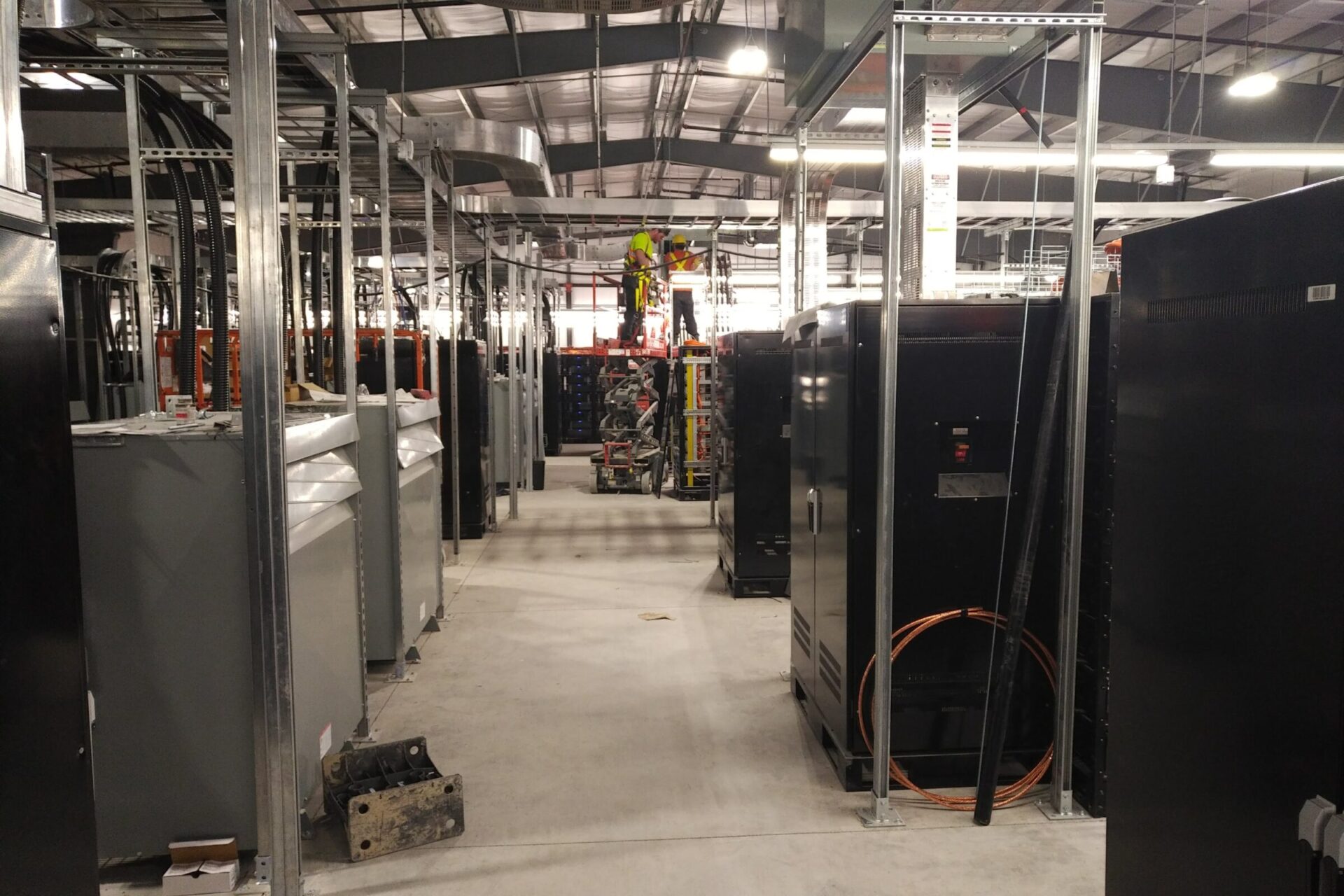 A person works on a scaffold surrounded by large machines and equipment in an industrial warehouse setting, with overhead lighting.