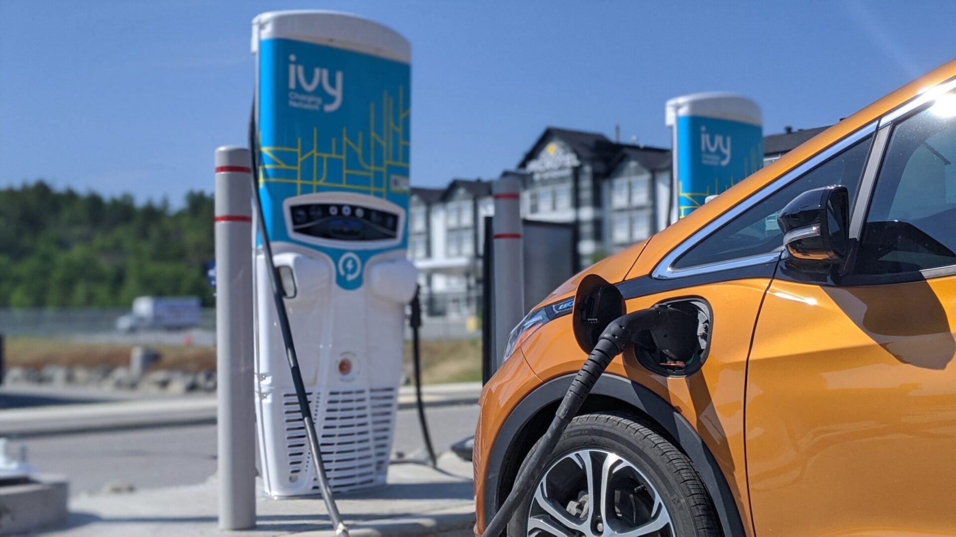 An orange electric vehicle is charging at an Ivy station, with residential buildings in the background under a clear blue sky.