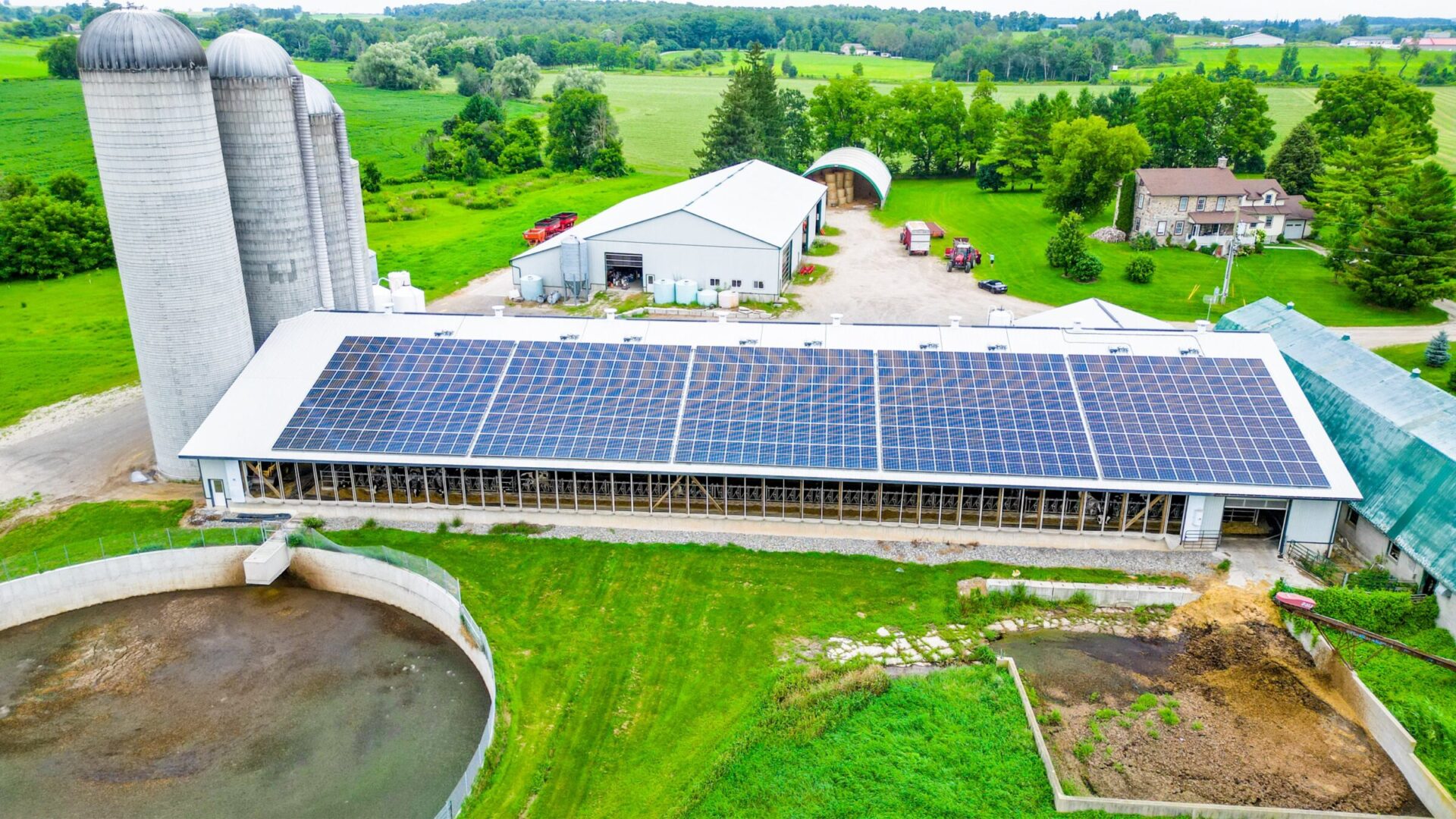 A farm with solar panels on a large barn, silos nearby, surrounded by greenery and fields under a clear sky.