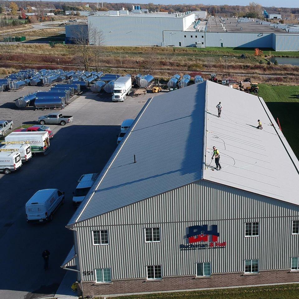 An industrial area with a large warehouse. Three people work on the roof. Surrounding vehicles and trailer trucks are visible.