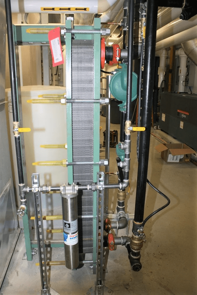 A mechanical room houses a large industrial heat exchanger with various pipes and valves, situated on a concrete floor in a utilitarian environment.