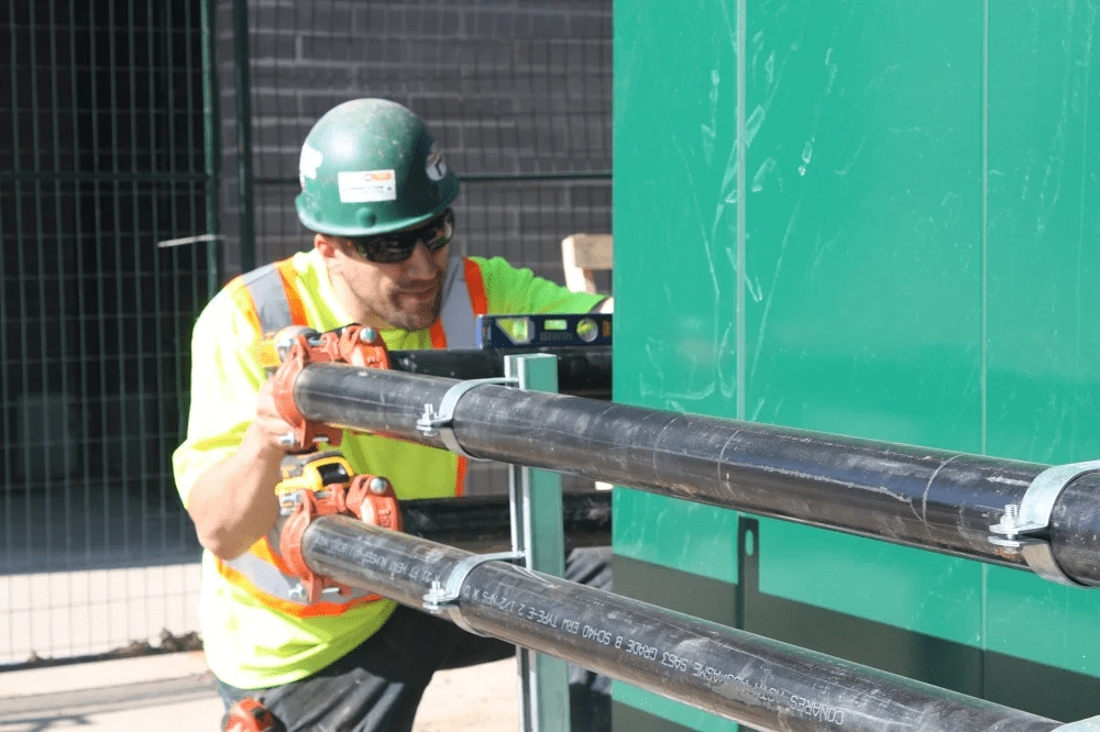 A person in safety gear and helmet is working with large pipes, using a level tool for alignment, next to a green structure.