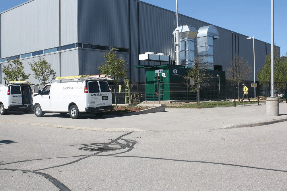 Two white utility vans parked outside a large industrial facility. A person stands nearby, with trees and clear blue sky in the background.