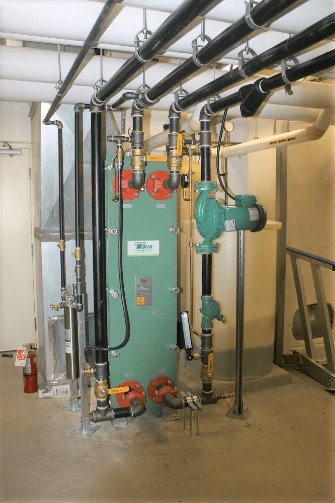 A mechanical room with interconnected pipes and valves, featuring a green heat exchanger. A fire extinguisher is mounted on the wall nearby.