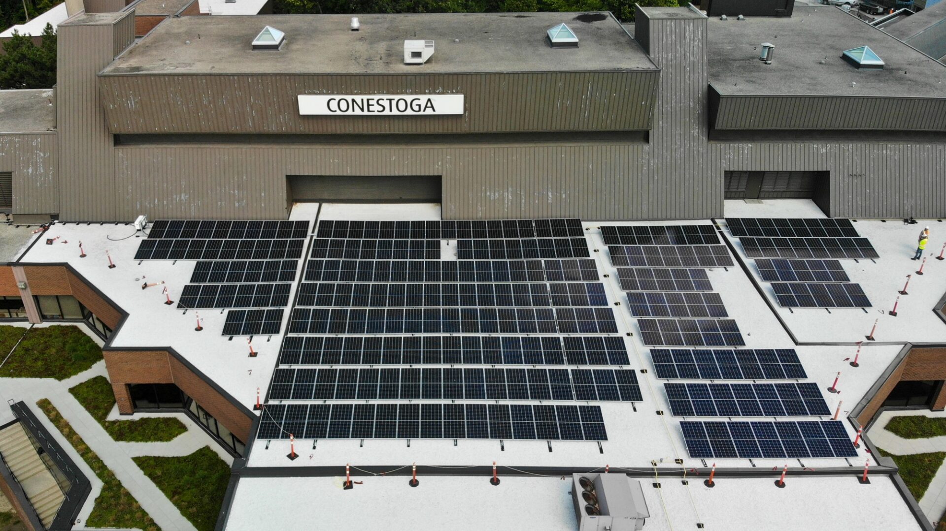 Aerial view of a building with "Conestoga" sign, featuring a large array of solar panels on the rooftop. No people visible.