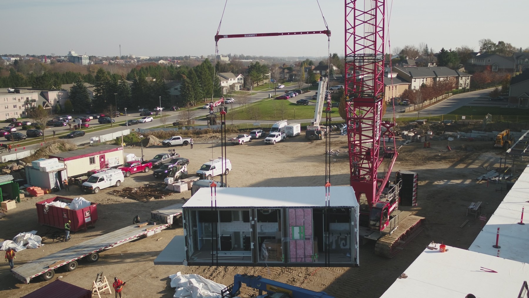 A large crane lifts a modular structure at a construction site, surrounded by vehicles and workers, under a clear sky.