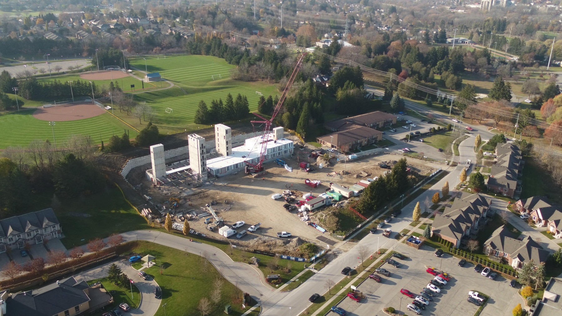 Aerial view of a construction site near residential area with sports fields in the background. Crane and vehicles are visible on the site.