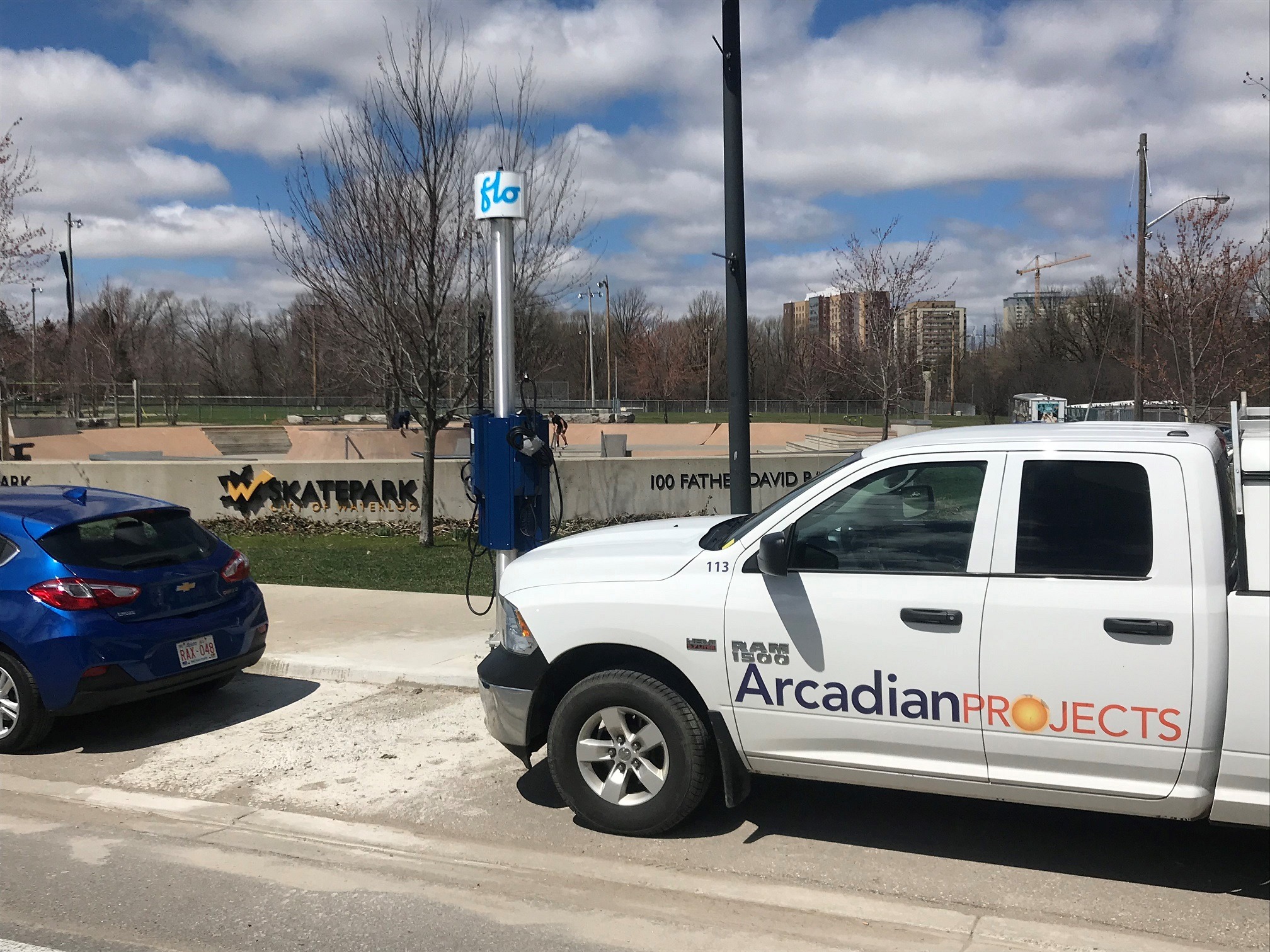 White utility truck and blue car parked near plug-in station at skatepark. Trees and buildings in the background. Clear sky with scattered clouds.