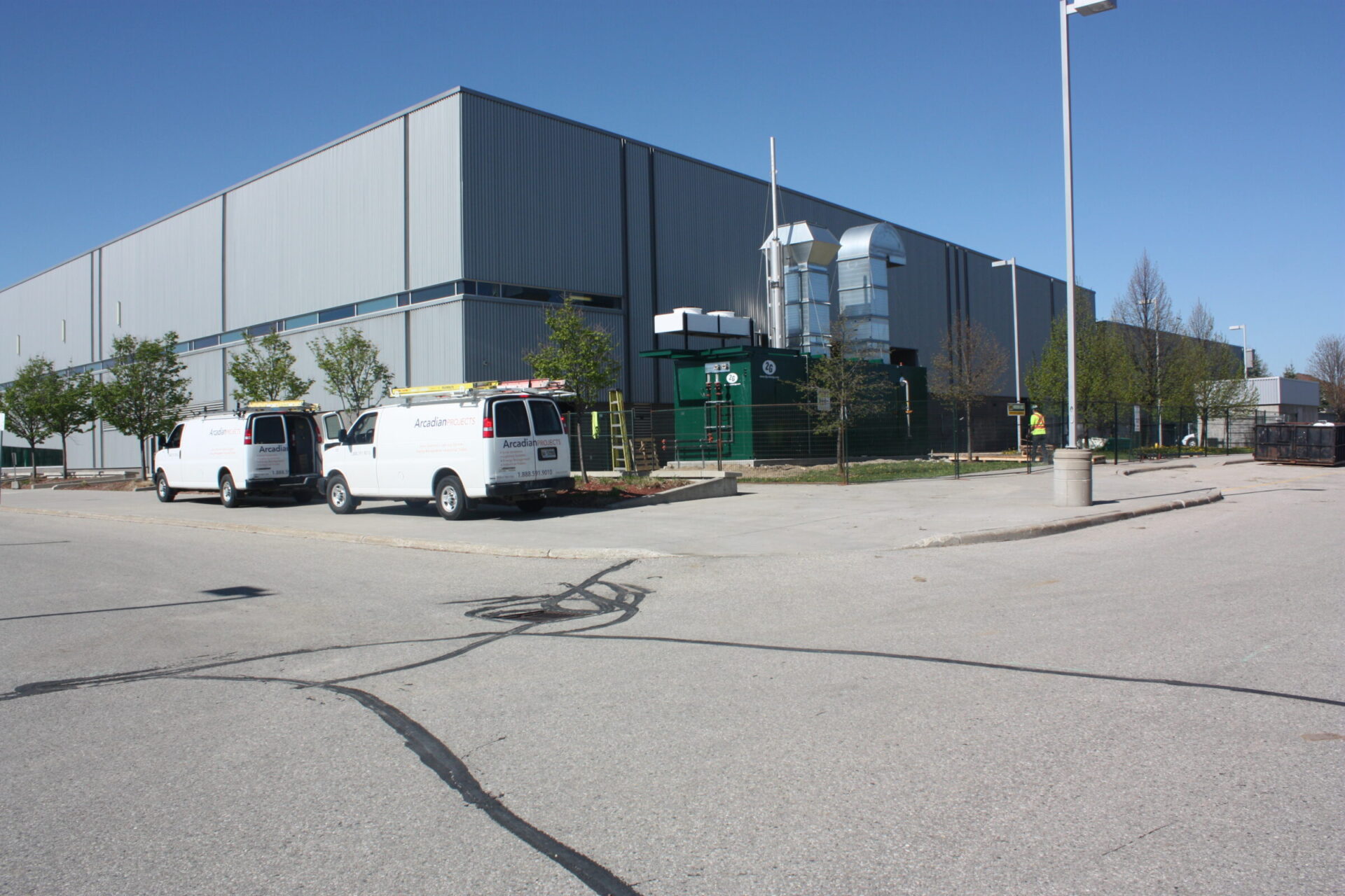 Industrial building with trees, two white vans parked outside. A person stands near the entrance. Clear sky above, empty paved area in foreground.