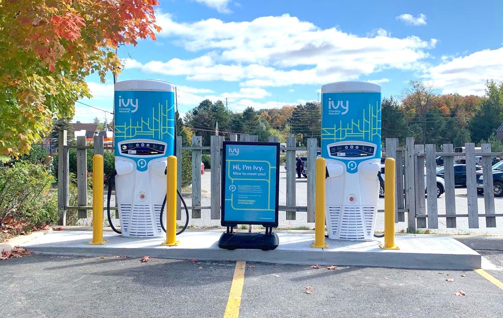 Electric vehicle charging station with two Ivy chargers in a parking lot. Surrounded by trees under a partly cloudy sky, with distant parked cars.