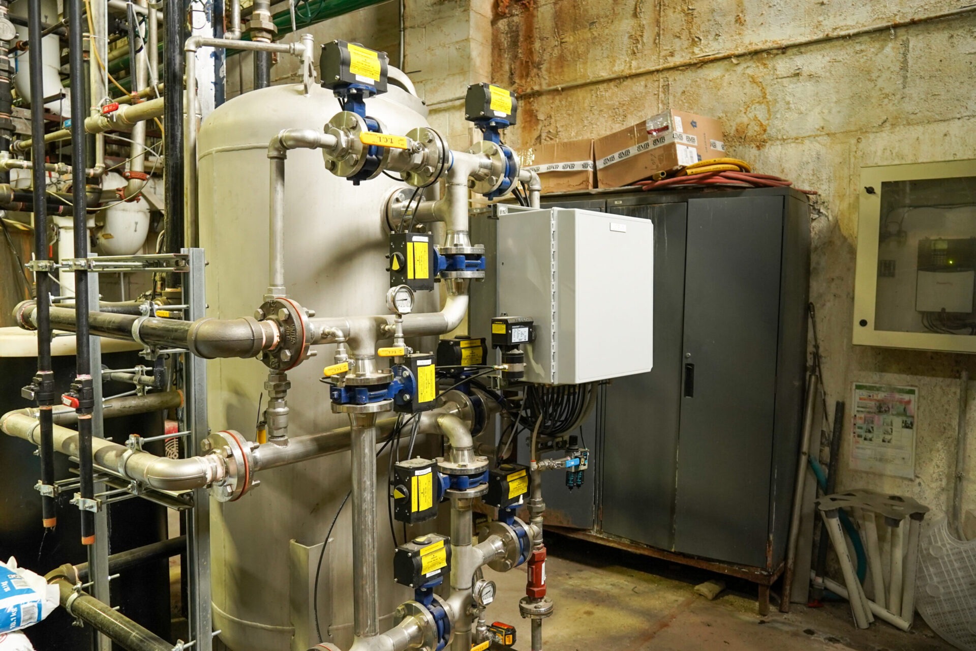 Industrial room featuring a complex system of pipes and valves attached to a large metal tank, with control panels and cabinets in the background.