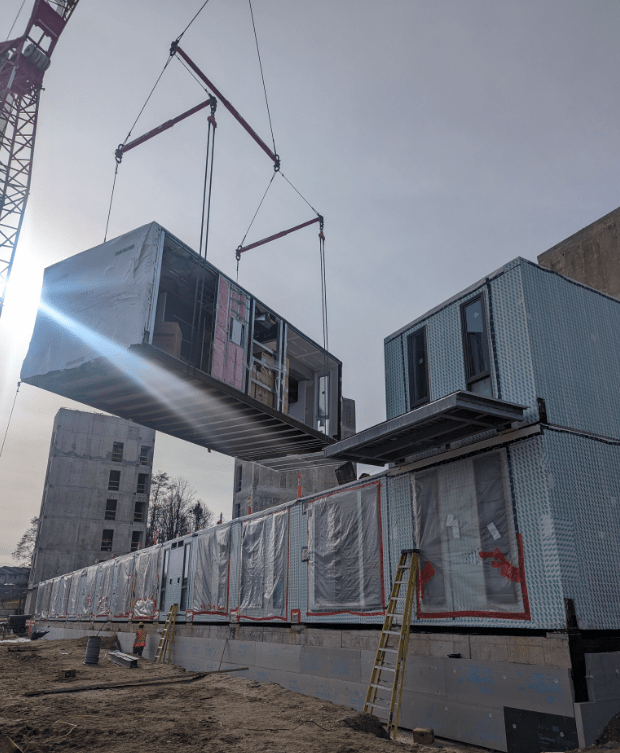 A modular building under construction, with a crane lifting a prefabricated section. Workers and equipment are visible on the construction site.