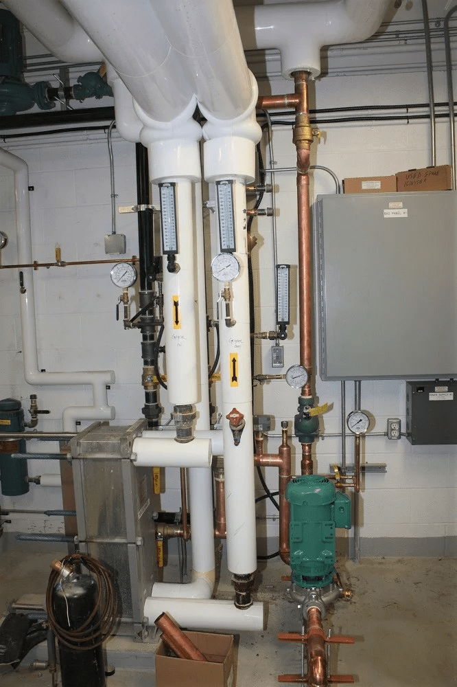 Industrial mechanical room with white insulated pipes, copper tubing, pressure gauges, and green pump. Equipment optimizes building systems. Concrete wall background.