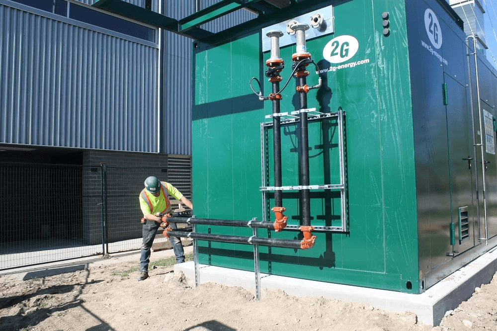 A person in safety gear works on a green industrial unit with pipes and valves outside a large metal building, under clear skies.