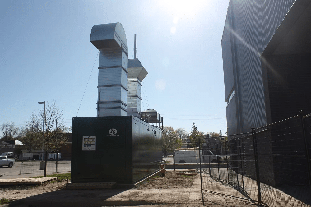 A large industrial air filtration system is outside a building, with a fenced area and trees in the background on a sunny day.