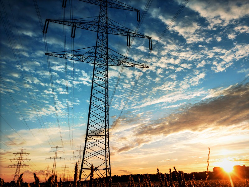 A row of electricity pylons stretches across a rural landscape at sunset, silhouetted against a vibrant sky with scattered clouds.