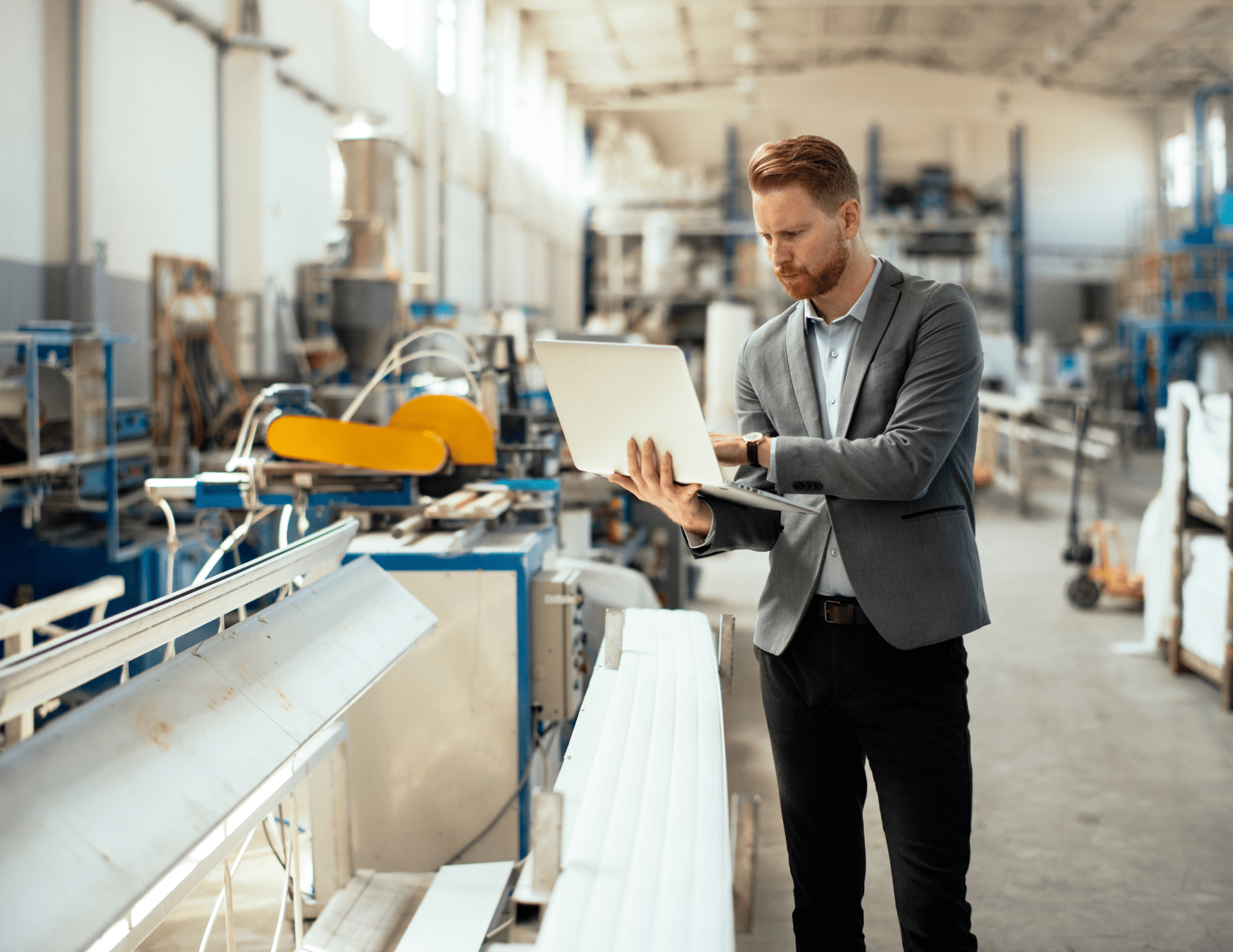 A person in a suit standing in an industrial workshop, holding a tablet, surrounded by machinery and equipment in a well-lit environment.