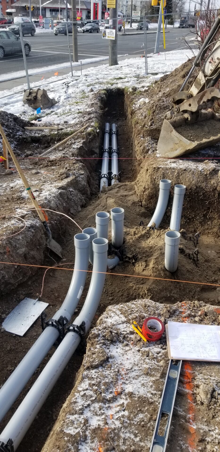 Construction site with trenches and installed pipes. Snow on the ground. Traffic and urban buildings in the background. Tools and plans visible on site.