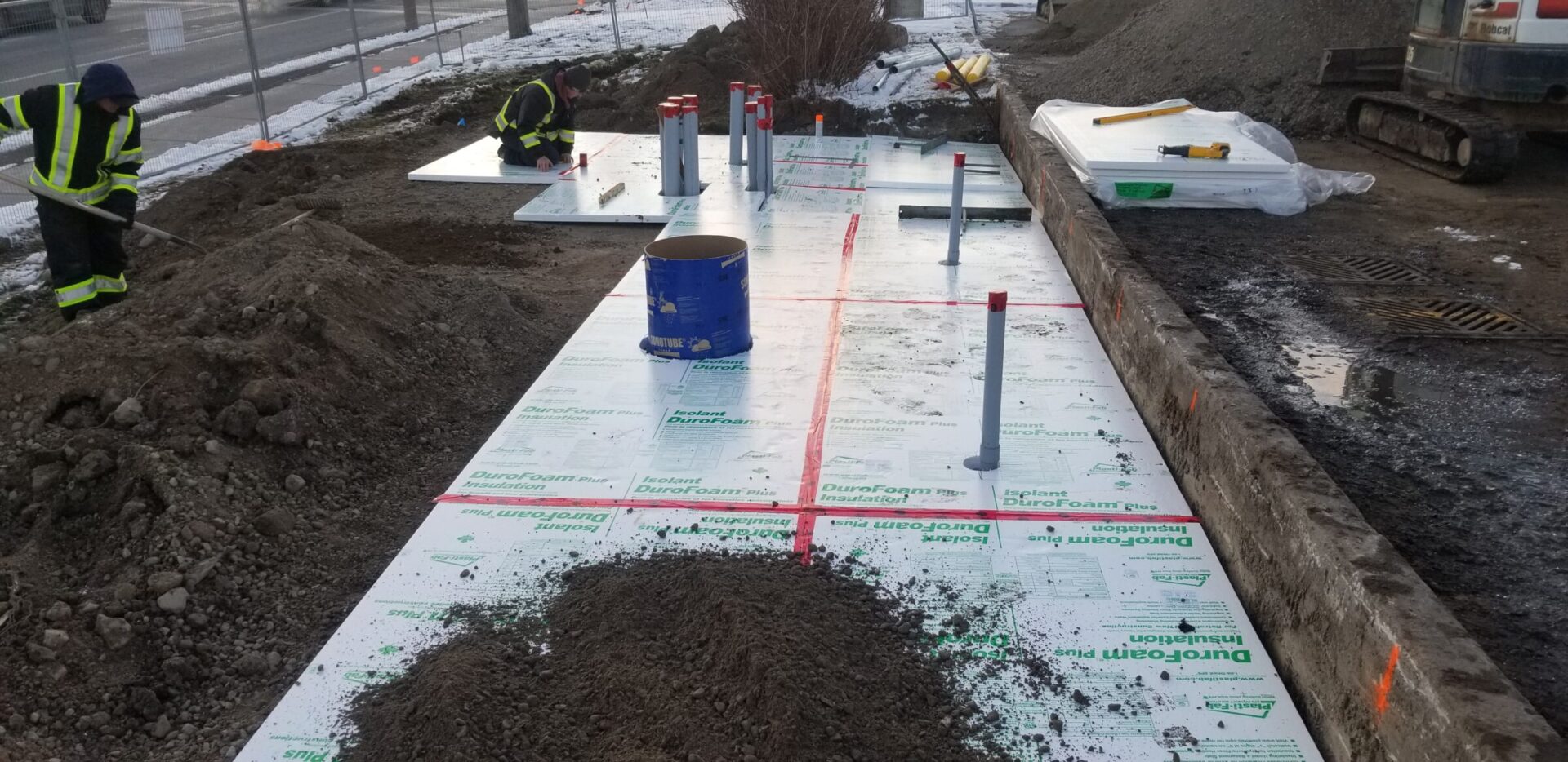 Two people in safety gear work on an insulated concrete slab, surrounded by construction equipment and materials, on a developing site.