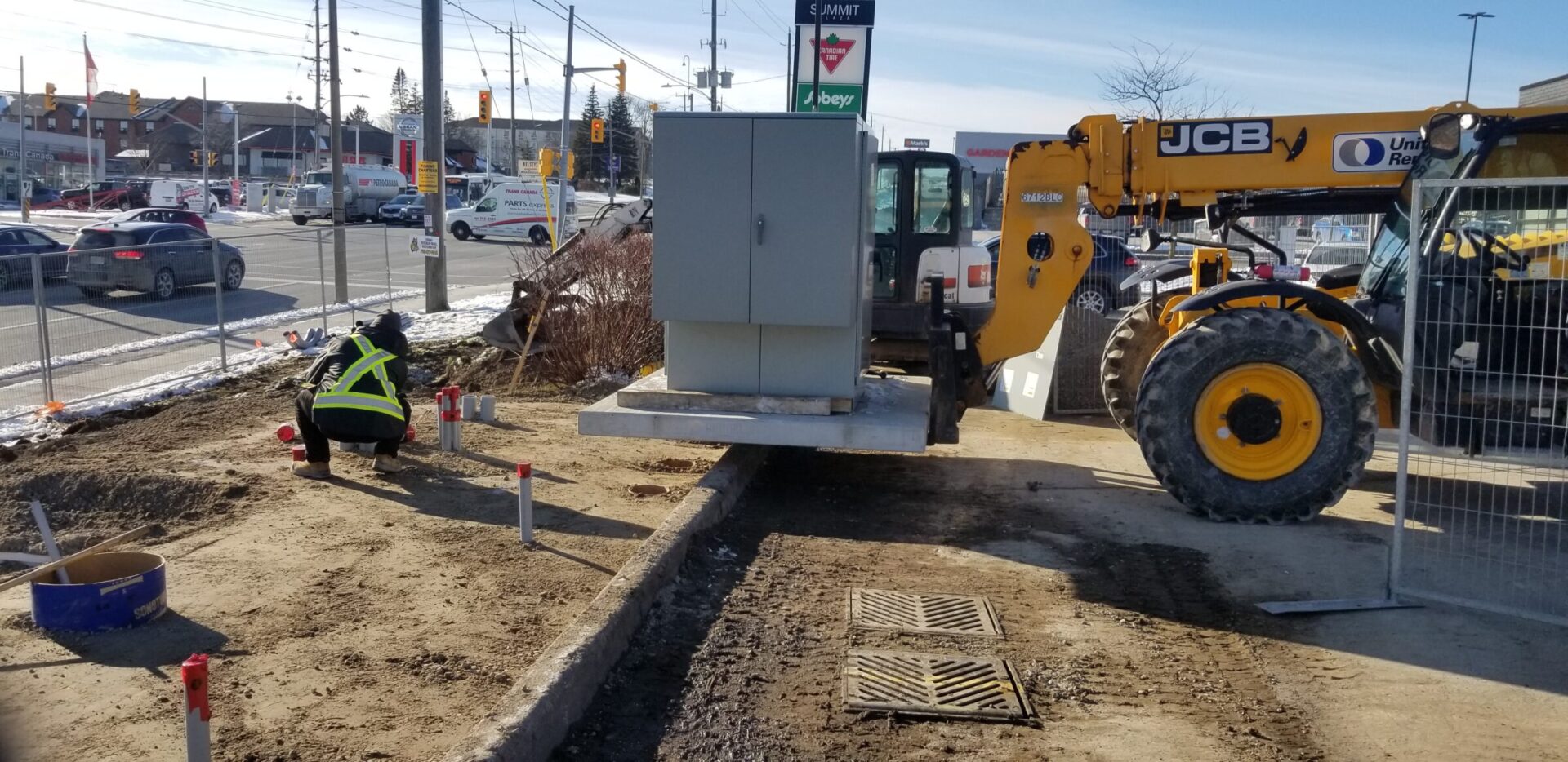 A construction site near a busy intersection, featuring a person working and a JCB machine. Urban buildings and traffic are visible in the background.