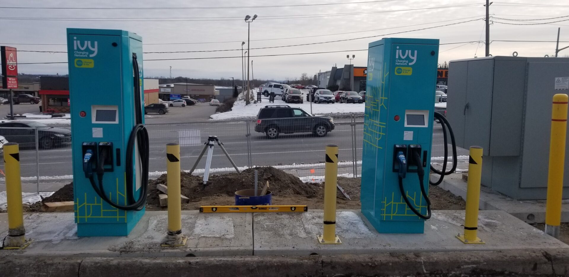 Two Ivy electric vehicle charging stations are situated in a snowy parking lot, with cars in the background and overcast sky above.