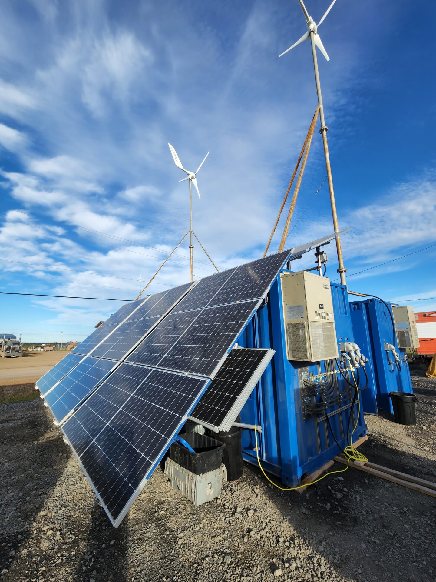 Solar panels and wind turbines stand against a clear blue sky. They're mounted on blue containers with electrical equipment, suggesting renewable energy use.