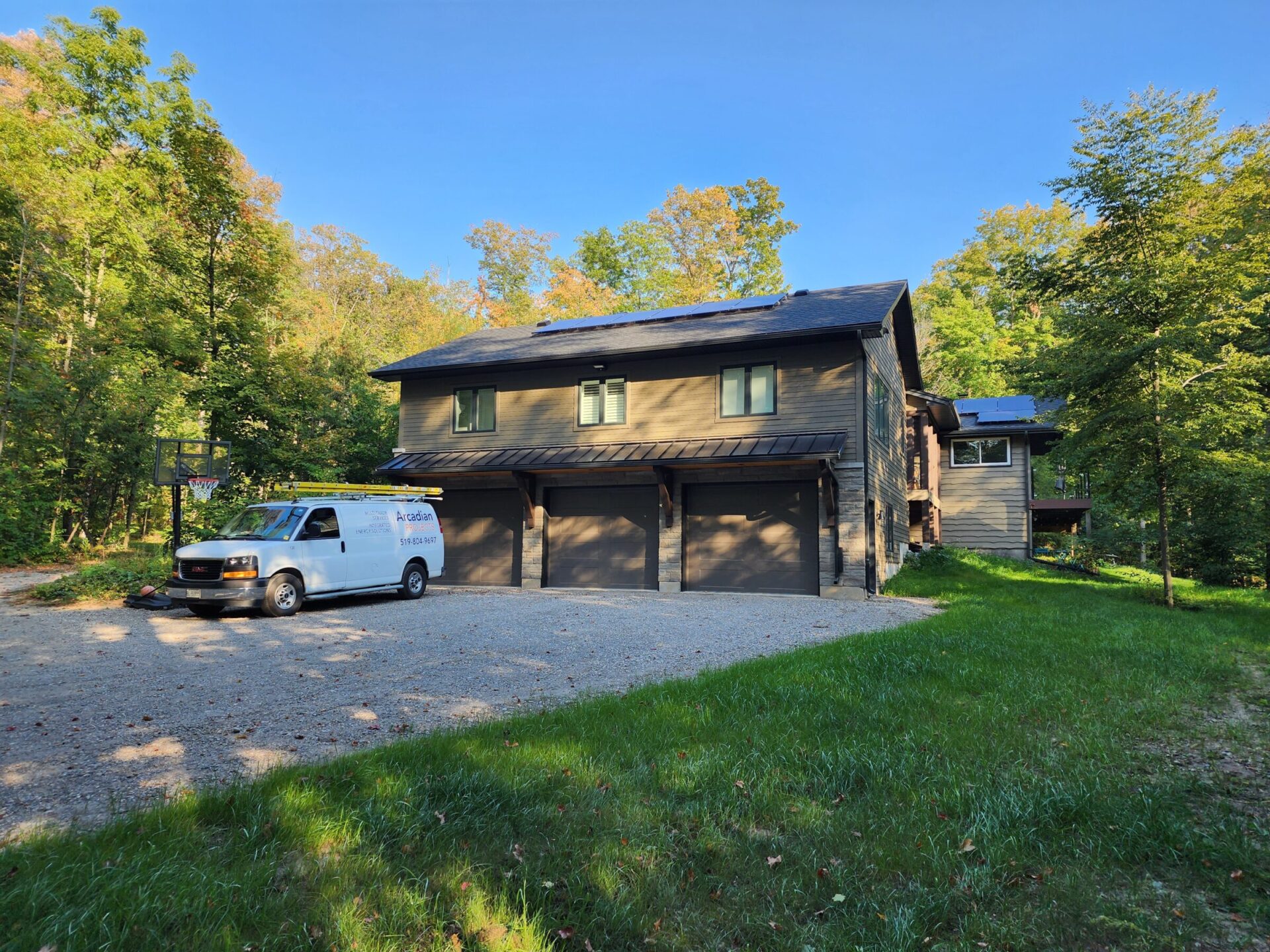 A white van is parked outside a large house surrounded by trees, under a clear blue sky, on a gravel driveway.