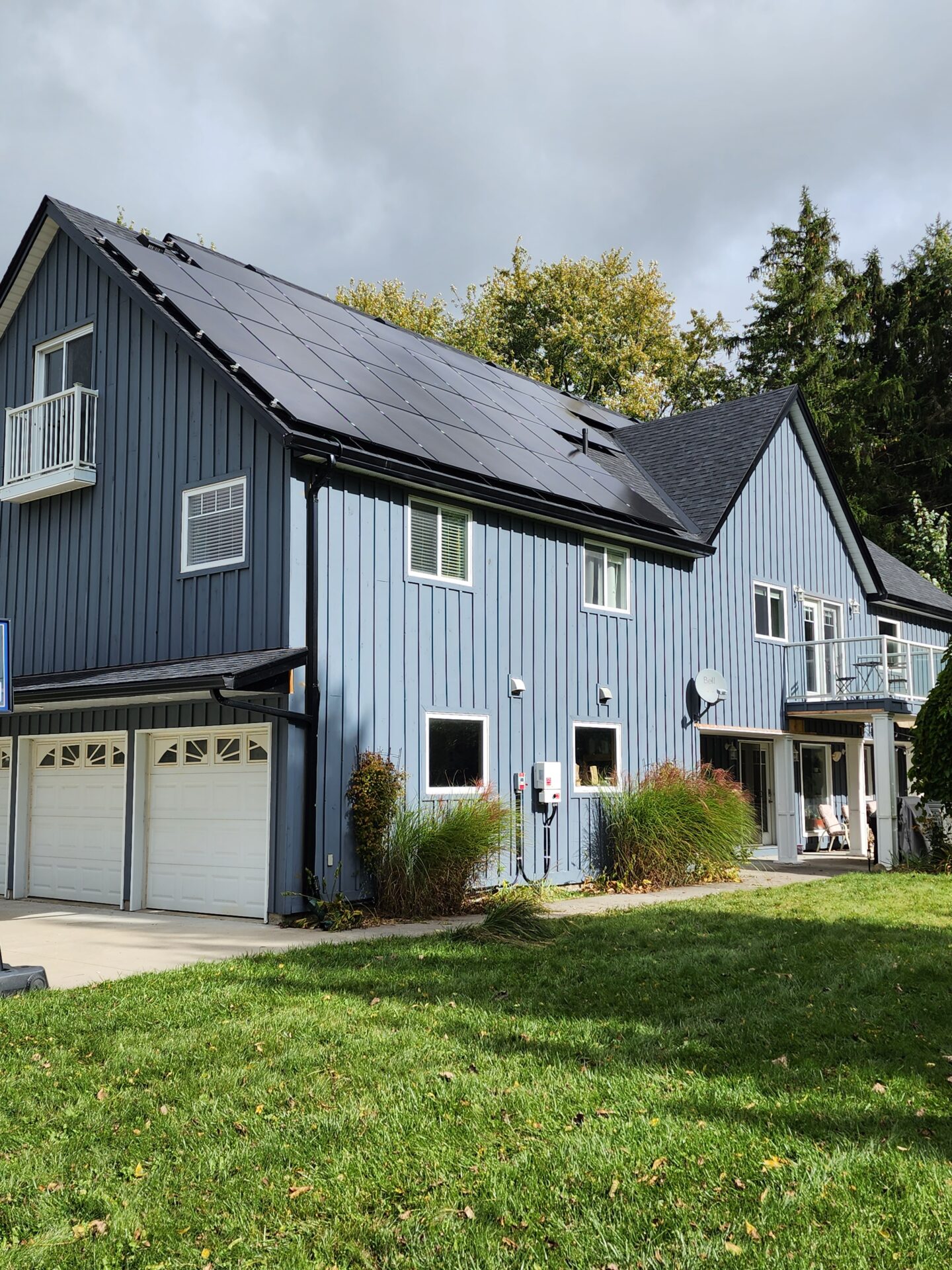 A blue house with solar panels and a two-car garage. A person is sitting on the porch, surrounded by greenery and trees.