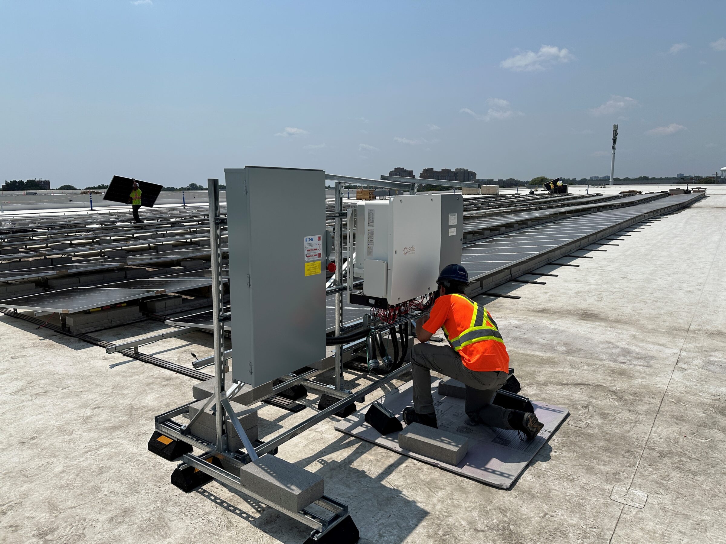 Person wearing a helmet and safety vest works on solar panel equipment on a large rooftop under a clear sky.