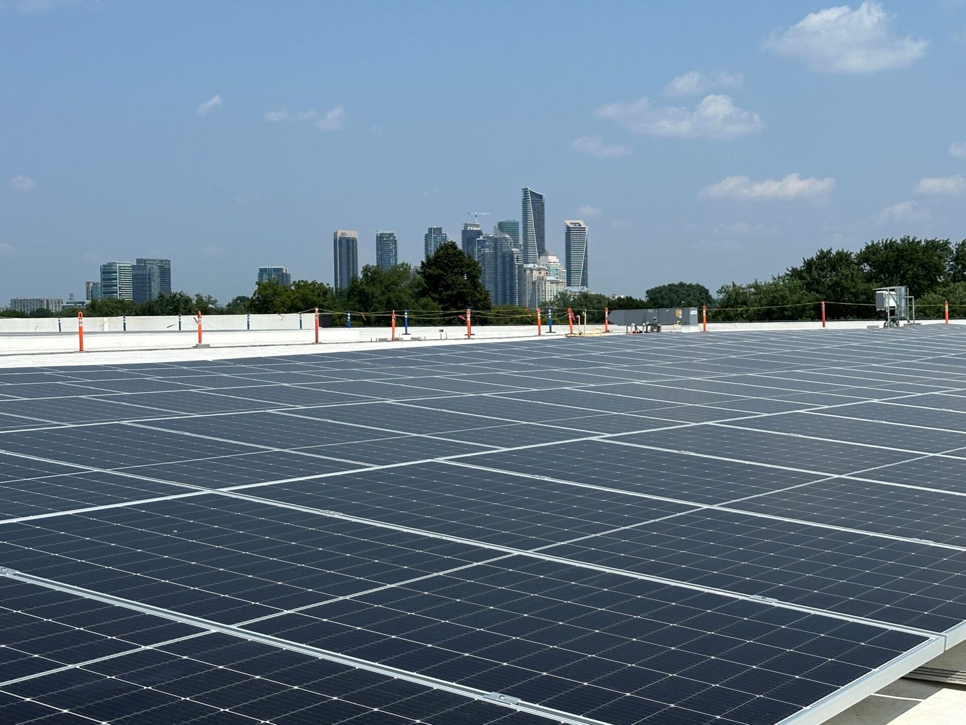 A large solar panel installation on a rooftop with a city skyline and trees in the background under a clear blue sky.