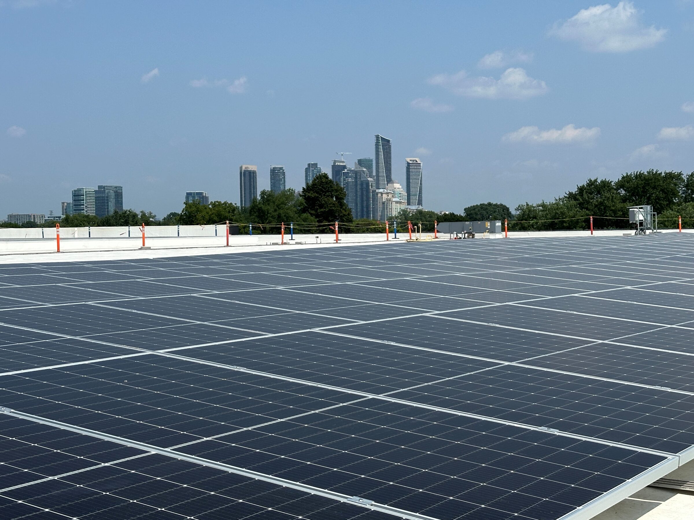 A large solar panel installation on a rooftop with a city skyline and trees in the background under a clear blue sky.