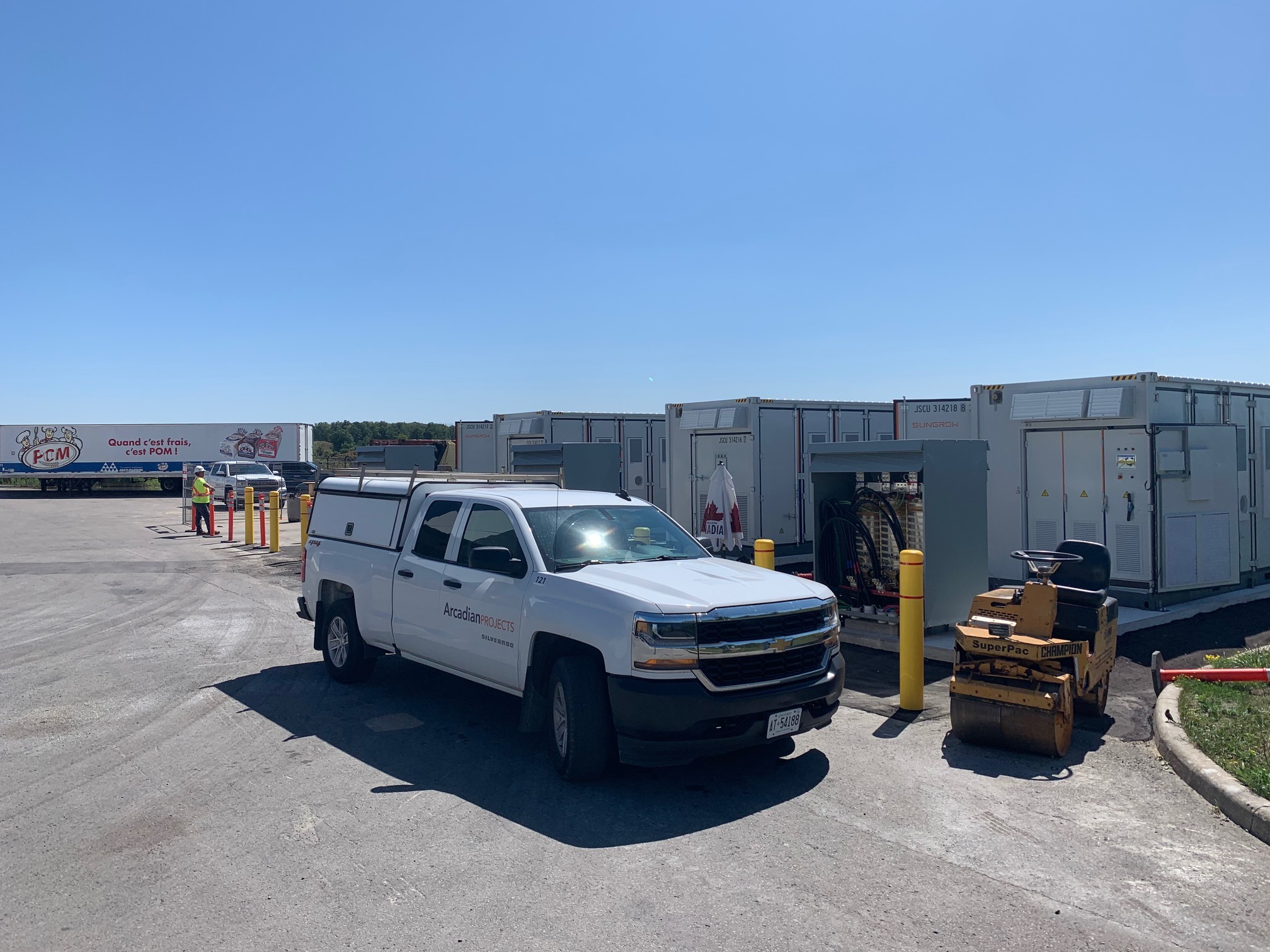 A white utility truck is parked near shipping containers and construction equipment under a clear sky. A sign is visible in the background.