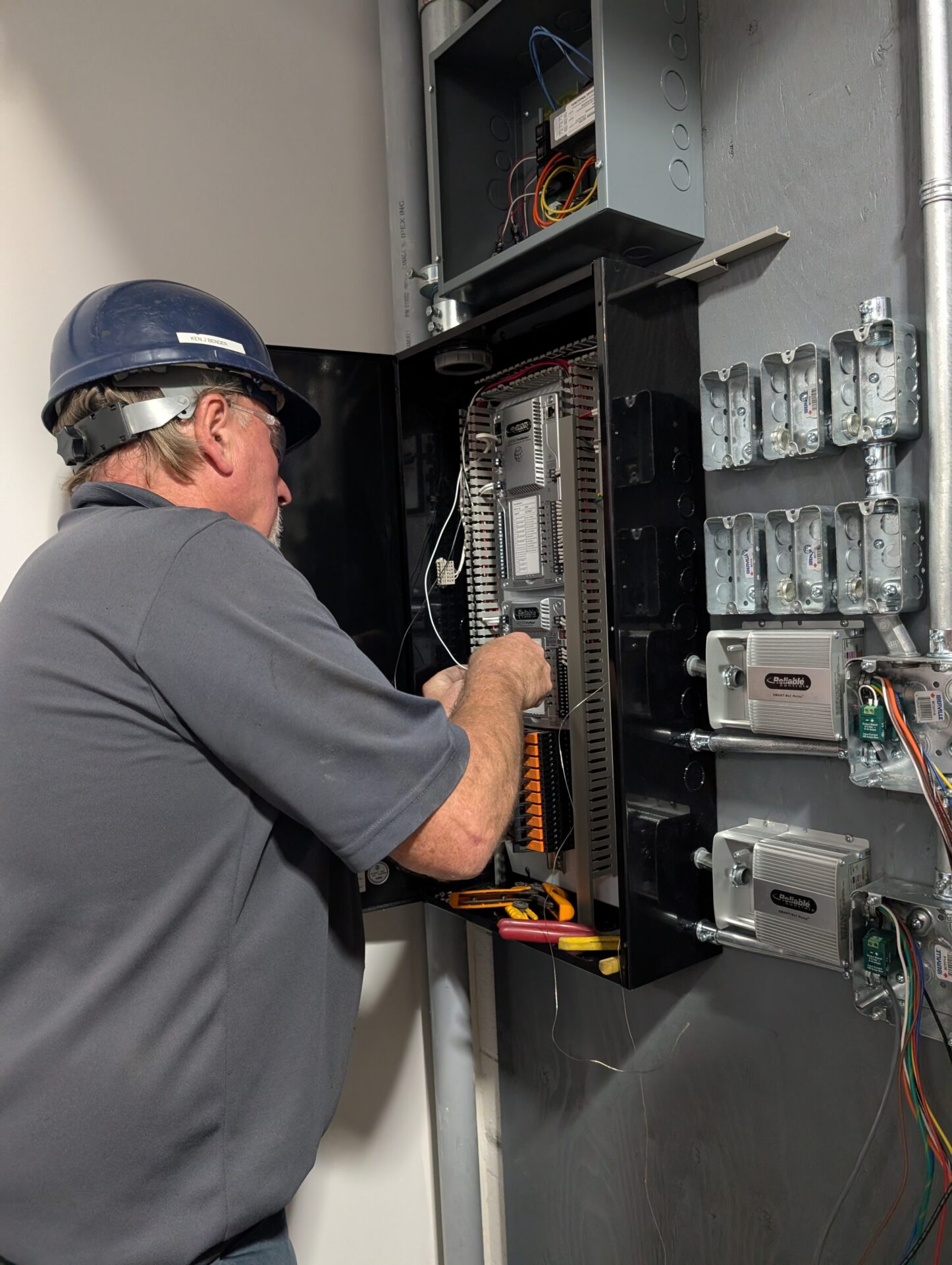 A person wearing a hard hat works on electrical panels in a utility room, surrounded by various wires and equipment.