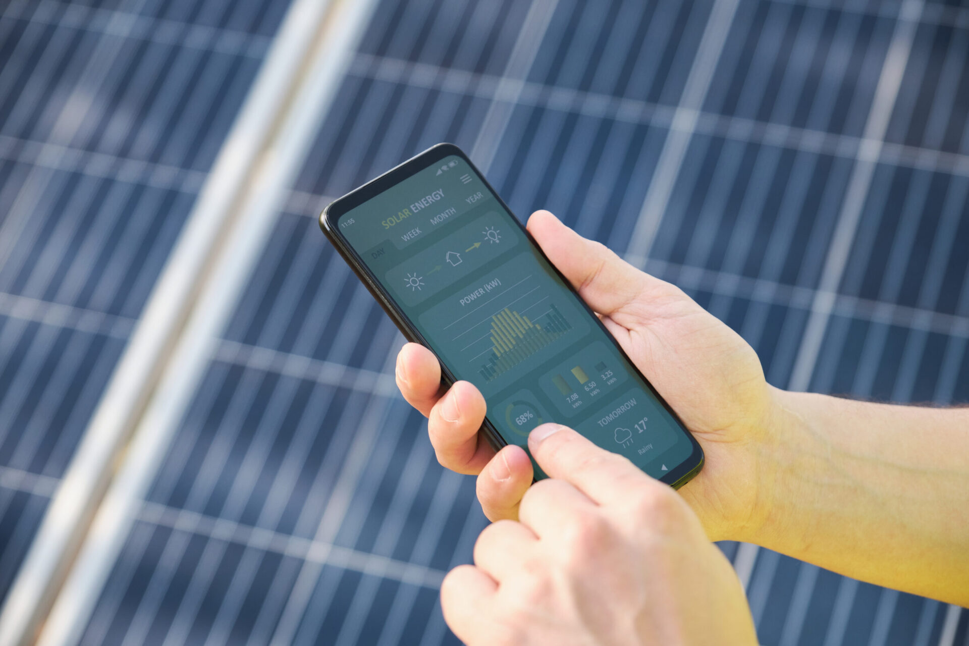 A person holds a smartphone displaying solar energy data, standing near large solar panels in bright sunlight.